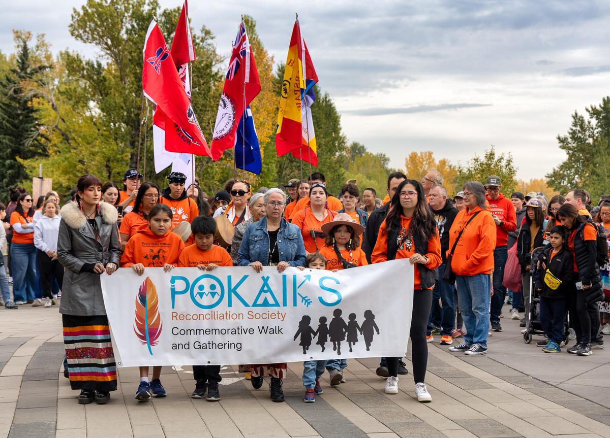 Families in orange shirts carry the Pokaiks Commemorative Walk and Gathering banner, with flags raised behind them.